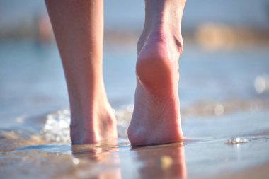 Close up of female feet walking barefoot on white grainy sand of golden beach on blue ocean water background.