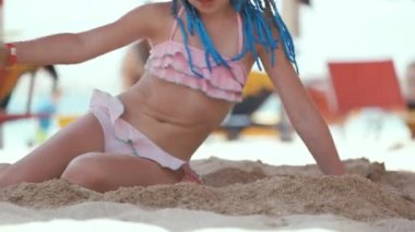 Happy child girl in swimsuit playing with sand under umbrella shadow during summer tropical vacations.