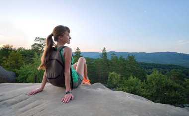 Sportive female hiker sitting alone taking a break on hillside trail. Lonely woman enjoying view of evening nature from rocky cliff on wilderness path. Active lifestyle concept.