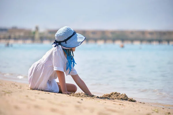 stock image Pretty child girl in big hat and white dress playing with wet sand on background of blue sky and clear ocean lagoon water.