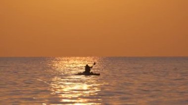 Sporcunun karanlık figürü gün batımında deniz suyunda kano teknesinde tek başına kürek çekiyor. Aktif ekstrem spor konsepti.