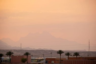 Uzak otel kompleksiyle gün batımı manzarası Mısır çölündeki karanlık dağ zirvelerine karşı.