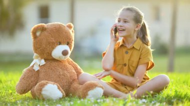 Güzel bir çocuk, arkadaşıyla birlikte yeşil çimlerin üzerinde oyuncak bir ayının yanında otururken tutkuyla cep telefonuna sarılıyor. Arkadaşlık değerleri kavramı.