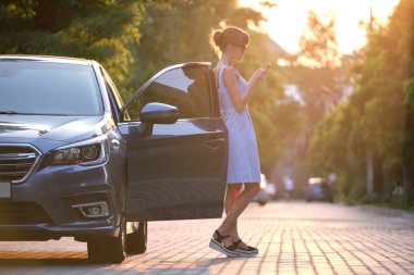 Aracının yanında duran genç bir kadın sürücü yazın bir şehir caddesinde cep telefonuna bakıyor..