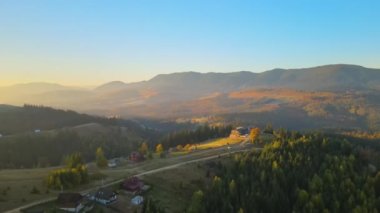 View from above of ukrainian Carpathian mountains with wooded hills and traditional village homes at autumnal sunset. Brightly illuminated pine woods with scattered local settlement houses.
