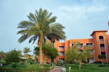 Fragment of red brick resort hotel exterior with palm trees under blue summer sky.