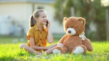 Dost canlısı bir çocuk, yeşil çimlerin üzerinde oyuncak bir ayıyla otururken arkadaşıyla telefonda mutlu bir sohbet ediyor. Arkadaşlık kavramı.