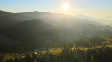 Aerial view of Carpathian mountain hills with small ukrainian village houses at sunset. Brightly illuminated pine woods with scattered local settlement homes at fall season.