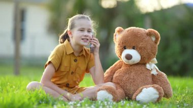 Mutlu kız çocuğu cep telefonuyla arkadaşına bir hikaye anlatıyor oyuncak ayısıyla dışarıda yeşil çimlerin üzerinde oturuyor. Arkadaşlık kavramı.