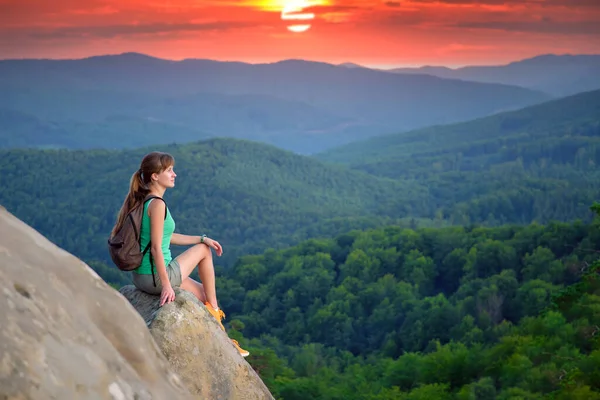 Tired hiker girl relaxing on rocky mountain top enjoying evening nature during travelling on wilderness trail. Lonely woman traveler traversing high hilltop route. Healthy lifestyle concept.