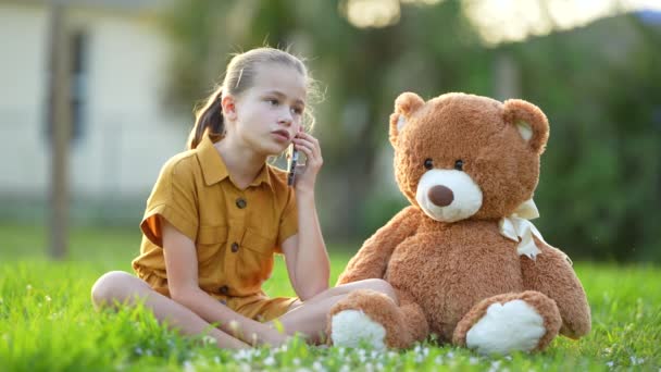 不快乐的小女孩在和朋友通电话的时候 和玩具熊坐在绿草草坪上 正在和朋友吵架 友谊问题的概念 — 图库视频影像