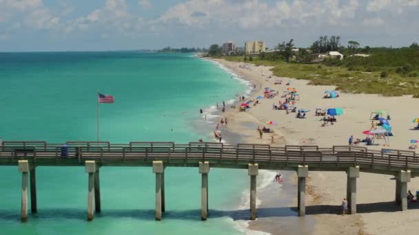 フロリダの有名なヴェネツィア釣り桟橋と砂浜 暖かい湾の水で入浴し 熱い太陽の下で日焼けを楽しんでいる多くの人の上を振るアメリカの旗 — ストック動画