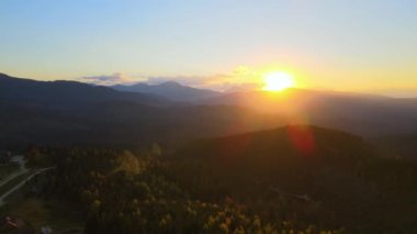 Aerial view of Carpathian mountain hills with small ukrainian village houses at sunset. Brightly illuminated pine woods with scattered local settlement homes at fall season.