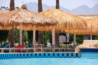 Boş güverte sandalyeleri, tropik tatil köylerinde havuz kenarında saman gölgesi şemsiyelerinin altında. Yaz tatilleri ve kaçış konsepti.