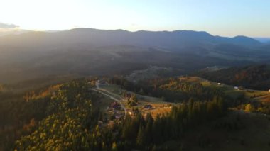 View from above of ukrainian Carpathian mountains with wooded hills and traditional village homes at autumnal sunset. Brightly illuminated pine woods with scattered local settlement houses.