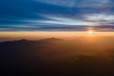 Gün batımında gün batımında parlak güneş ışınlarıyla karanlık dağ tepelerinin havadan görünüşü. Akşamları sisli tepeler ve sisli vadiler.