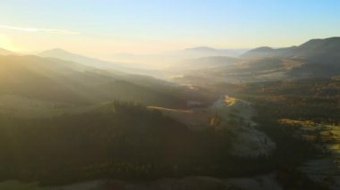 Aerial view of Carpathian mountain hills with small Ukrainian village houses at sunset. Brightly illuminated pine woods with scattered local settlement homes at fall season.