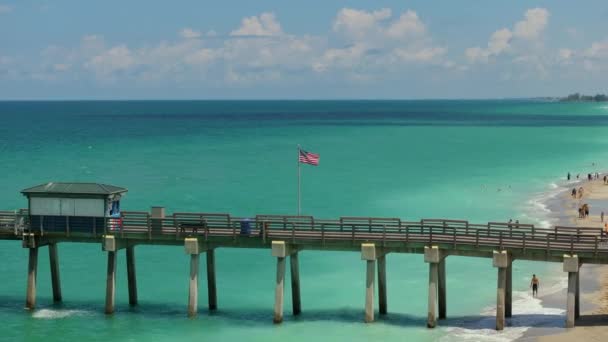 フロリダ州のヴェネツィア フィッシング ピア近くの砂浜に手を振っているアメリカ国旗 休暇中の水泳や日焼けを楽しむ観光客 海辺の夏のアクティビティ — ストック動画