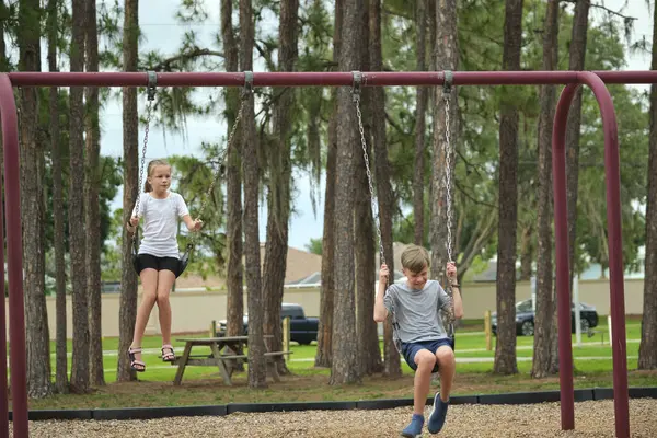 Two young teenage children, girl and boy playing on swings together outdoors on bright sunny vacations day.