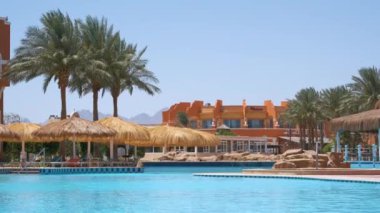 Boş güverte sandalyeleri, tropik tatil köylerinde havuz kenarında saman gölgesi şemsiyelerinin altında. Yaz tatilleri ve kaçış konsepti.