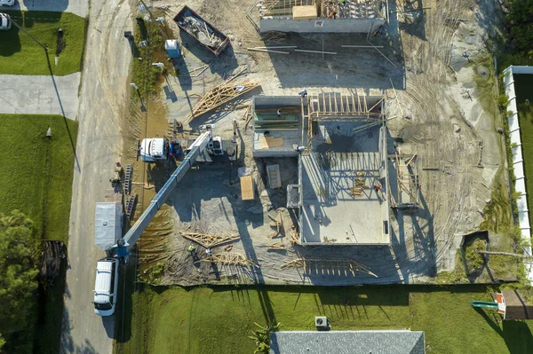 Aerial view of lifting crane and builders working on unfinished residential house with wooden roof frame structure under construction in Florida suburban area.