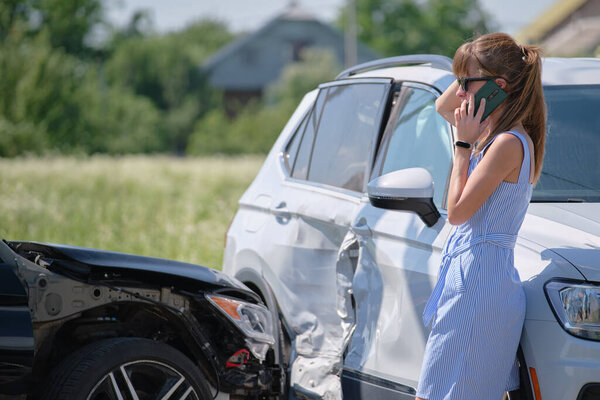 Stressed driver talking on sellphone on roadside near her smashed vehicle calling for emergency service help after car accident. Road safety and insurance concept.