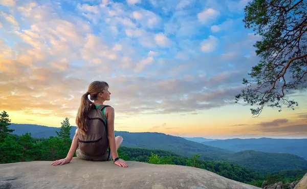 Young woman hiker sitting alone on rocky mountain enjoying view of evening nature on wilderness trail. Active way of life concept.