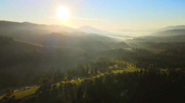 Wooded hills of Carpathian mountains in Ukraine with small traditional village houses at sunset. Brightly illuminated with setting sun evergreen woods with scattered local homes at fall season.