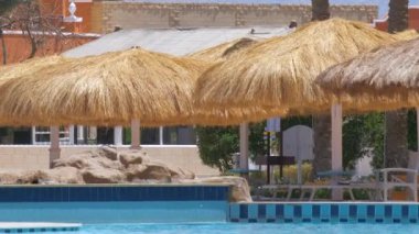 Boş güverte sandalyeleri, tropik tatil köylerinde havuz kenarında saman gölgesi şemsiyelerinin altında. Yaz tatilleri ve kaçış konsepti.