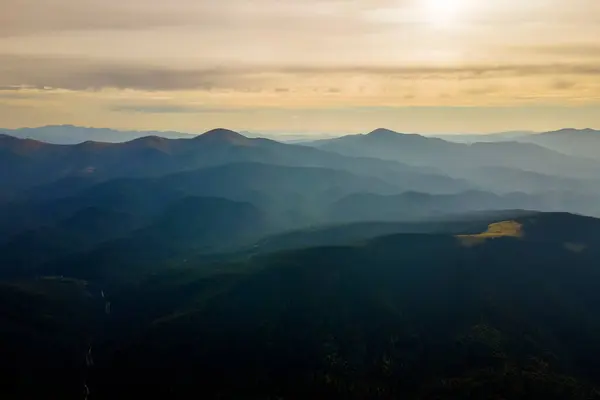 Aerial view of dark mountain hills with bright sunrays of setting sun at sunset. Hazy peaks and misty valleys in evening.