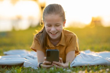 Child girl texting with her friend on cellphone outdoors in summer park.