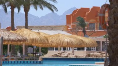 Boş güverte sandalyeleri, tropik tatil köylerinde havuz kenarında saman gölgesi şemsiyelerinin altında. Yaz tatilleri ve kaçış konsepti.