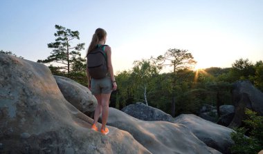 Sportive woman walking alone on hillside trail. Female hiker enjoying view of evening nature from rocky cliff on wilderness path. Active lifestyle concept.