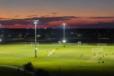 Halk parkındaki aydınlık stadyumda futbol oynayan sporcular.