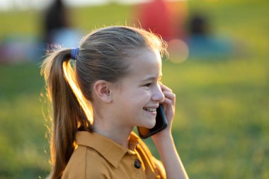 Child girl talking with her friend on cellphone outdoors in summer park.