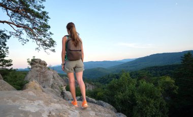 Sportive woman walking alone on hillside trail. Female hiker enjoying view of evening nature from rocky cliff on wilderness path. Active lifestyle concept.