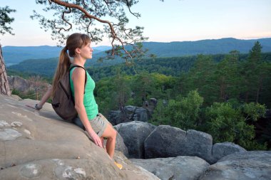 Sportive female hiker sitting alone taking a break on hillside trail. Lonely woman enjoying view of evening nature from rocky cliff on wilderness path. Active lifestyle concept.