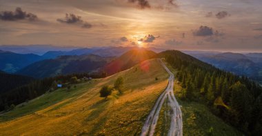 Vahşi dağlarda renkli günbatımının havadan görünüşü. Çam ormanlarından geçen karanlık toprak yol parlak batan güneşle aydınlanıyor. Güzel doğa manzarası.