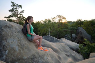 Sportive female hiker sitting alone taking a break on hillside trail. Lonely woman enjoying view of evening nature from rocky cliff on wilderness path. Active lifestyle concept.