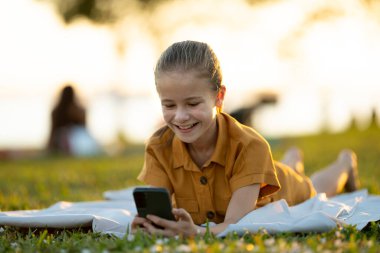 Mutlu kız çocuğu yaz parkında cep telefonuyla video oyunu oynuyor..