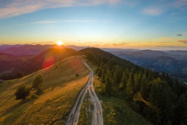 Tepenin tepesinde dönen toprak yol parlak batan güneşle aydınlanıyor. Vahşi dağlarda renkli günbatımının havadan görünüşü. Güzel doğa manzarası.