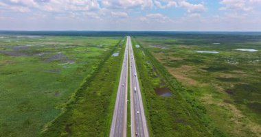 Florida otoyolunun hava manzarası mavi gökyüzünün altında hareket eden araçlarla doğal sulak araziyi kesiyor.
