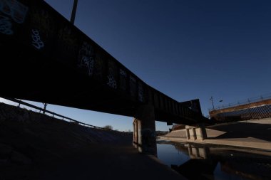 A dark railway bridge with graffiti spans a shallow river near a border fence topped with barbed wire
