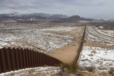 An aerial perspective shows a long section of the border wall extending across a snowy, arid landscape with scattered vegetation