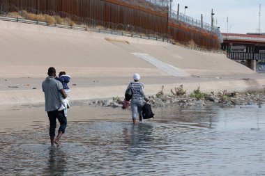 Bir adam sığ bir nehirde yürürken bir çocuk taşır ve ardından bir kadın bir çanta taşır.