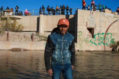 Kot ceket giyen ve turuncu şapkalı bir adam sığ, dalgalı bir nehirde yürüyor.