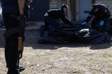 Üç birey, siyah bir ceset torbasının yanındaki toprak ve yamalı çimlerin üzerinde görünür.