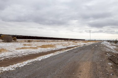 A dirt road runs parallel to a long section of the U.S.-Mexico border wall in a flat, arid landscape dotted with sparse vegetation and patches of snow, under a heavy, overcast sky