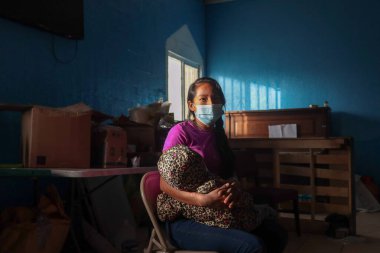 Sınır dışı edilmiş, mor tişörtlü, koyu mavi kotlu ve açık mavi cerrahi maskeli bir kadın Ciudad Jurez, Chihuahua 'da bir barınağın içindeki metal bir sandalyede oturup çocuğunu kucaklıyor.