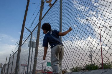 Koyu renk gömlekli ve açık renk pantolonlu genç bir çocuk Ciudad Jurez 'de bir barınağın içindeki uzun zincirli çitin yanında dikilip yukarıya bakıyor.
